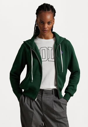Felpa verde con zip, tasca a marsupio, coulisse bianche e logo ricamato. Indossata sopra una maglietta bianca con testo in grassetto nero. Pantaloni grigi.