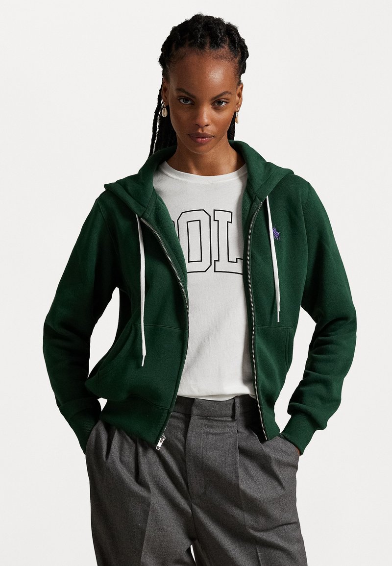 Grøn zip-up hoodie med en kængurulomme, hvide snore og broderet logo. Bæres over en hvid t-shirt med fed sort tekst. Grå bukser.