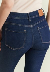 Jeans de mezclilla azul oscuro con un diseño de cintura alta, que presentan costuras naranjas en contraste y dos bolsillos traseros. Silueta ligeramente ajustada.