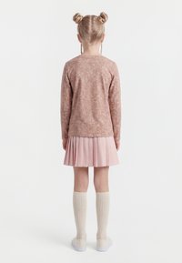 Mladá dívka s blond vlasy staženými do dvou drdolů, oblečená v růžovém svetru, plisované růžové sukni, bílých ponožkách po kolena a bílých botách, stojící zády.