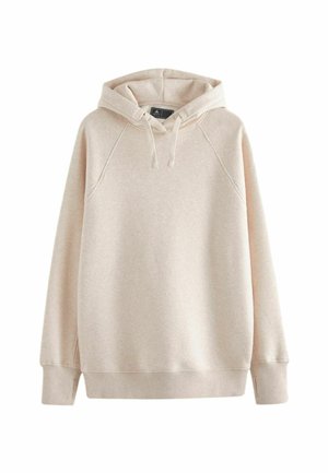 Personne portant un sweat à capuche beige clair avec des cordons et un pantalon marron foncé, debout devant un fond gris uni.