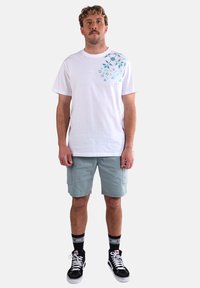 T-shirt en coton blanc avec un imprimé floral turquoise sur l'épaule gauche, associé à un short cargo gris clair, des baskets noir et blanc, et des chaussettes rayées.