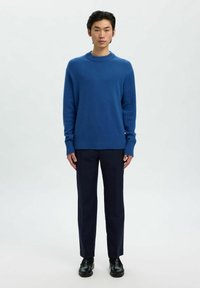 Joven de pie contra un fondo blanco, vistiendo un suéter azul, pantalones oscuros y zapatos de cuero negro, mirando hacia adelante.