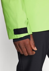 Gros plan d'une main à la peau foncée portant une veste vert néon avec une bande Velcro noire au poignet et un pantalon noir.
