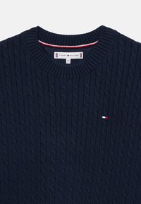 Marineblaue Kabelstrickpullover mit einem gerippten Rundhalsausschnitt, strukturiertem Muster und einem kleinen Logo mit roten und weißen Akzenten auf der Brust.