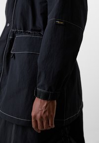 Nærbilde av en person som har på seg en svart Cordura-jakke med hvite stikninger og borrelås-lukking ved håndleddet.
