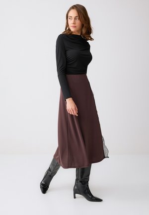 Femme portant un haut à manches longues noir, une jupe midi marron avec une bordure en dentelle, et des bottes à talons noires, debout devant un fond blanc uni.