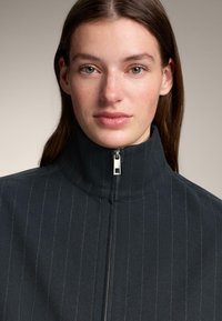 Fekete, cipzáros dzseki magas gallérral, finom tűcsíkos mintával, sima anyagból készült, fém cipzár díszítéssel.