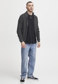 Cardigan cu fermoar de culoare gri închis, din țesătură texturată, purtat peste o cămașă neagră. Asortat cu blugi albastri deschis și adidași negri.