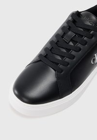 Adidași negri din piele cu șireți negri și talpă albă, cu branding Calvin Klein pe lateral și pe limbă.