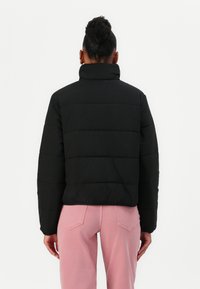 Svart puffjacka med hög krage och horisontell kviltad design. Bärs över rosa jeans, vilket framhäver en kontrasterande färgschema.