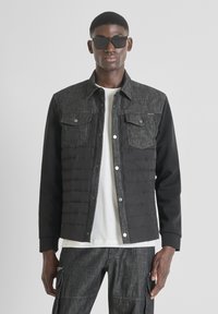 Giacca nera e grigia con una parte superiore simile al denim, sezione inferiore imbottita nera, tasche frontali e bottoni a scatto metallici. Indossata sopra una camicia bianca.