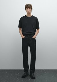 Sort bomulds T-shirt og sorte denim jeans. Modellen står med hænderne i lommerne. Sorte lædersko, minimalistisk design, glat tekstur.