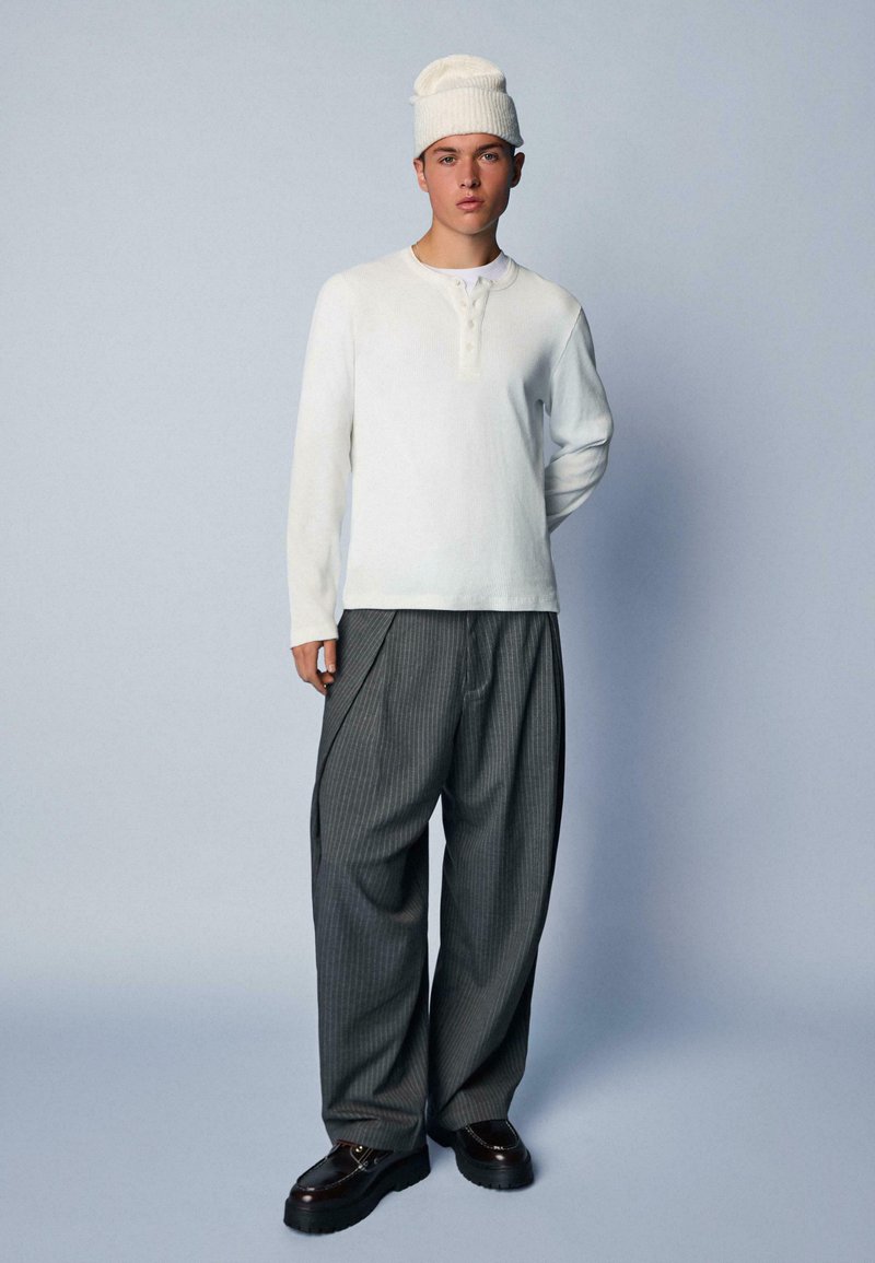 Langärmliges weißes Henley-Shirt, graue, gestreifte weite Hosen und eine gerippte weiße Mütze. Schwarze Plateauschuhe runden das Outfit ab.