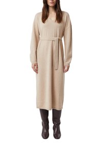 Beżowa dzianinowa sukienka kopertowa z dekoltem w serek, długimi rękawami i paskiem do wiązania w talii. Dół sukienki i mankiety rękawów mają prążkowaną teksturę. Połączona z brązowymi botkami.