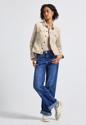 Une femme se tient avec les jambes croisées, portant une veste beige à boutons, un haut clair, un jean bleu retroussé et des baskets blanches devant un fond uni.