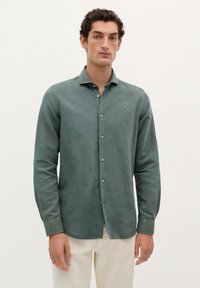 Camisa verde de manga larga hecha de tela texturizada, con un cuello abotonado y botones en la parte frontal, y un sutil logo en el pecho.
