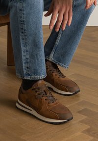 Baskets en cuir marron avec une finition texturée, dotées de semelles blanches et de lacets noirs, associées à un jean bleu clair légèrement fuselé.