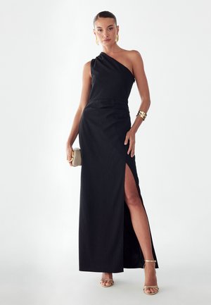 Kobieta w czarnej wieczorowej sukni na jedno ramię z rozcięciem do uda, złotych sandałach, bransoletce, kolczykach i trzymająca beżową kopertówkę.