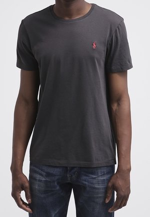 Czarna bawełniana koszulka z krótkim rękawem i okrągłym dekoltem. Posiada małe czerwone logo na lewej piersi. Noszona z niebieskimi dżinsami.