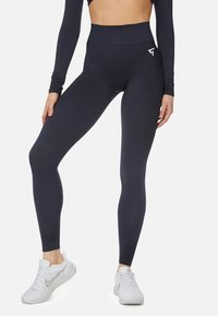 Navy high-waisted leggings in a stretchy material with textured accents. Paired with white athletic shoes, featuring a logo on the waistband.