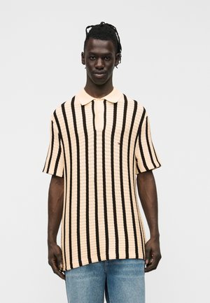 Uomo con pelle scura e capelli intrecciati che indossa una polo beige con righe verticali nere e jeans blu, in piedi davanti a uno sfondo bianco.