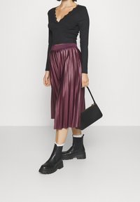 Top nero a maniche lunghe con collo scallop, abbinato a una gonna midi plissettata borgogna. La modella indossa stivali neri chunky e tiene una piccola borsa nera.