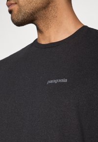 Čierne bavlnené tričko s dlhým rukávom, okrúhlym výstrihom a jemným sivým logom "Patagonia" na ľavej strane hrudníka. Hladká textúra.