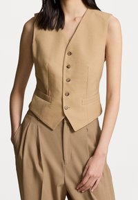 Béžová vesta bez rukávů vyrobená z texturované látky, s výstřihem do V, pěti knoflíky vpředu a dvěma bočními kapsami, s přiléhavým střihem.