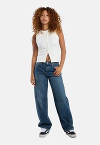 White sleeveless top with a button front, paired with loose blue denim jeans. Model wears black and white sneakers.