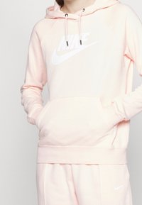 Ljusrosa Nike-huvtröja med en stor vit logotyp, snörning och en framficka. Tyget verkar mjukt med en bekväm passform.