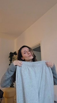 Saia cinza claro com cintura elástica, feita de um tecido texturizado, sustentada por uma pessoa a usar um suéter cinza num interior neutro.