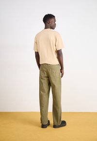 Beige cotton t-shirt, olive green work pants with rear pockets, black shoes, against a neutral backdrop with a yellow floor.