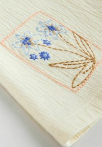 Tissu crème avec broderie florale bleue et marron dans un cadre rectangulaire, présentant une texture subtile et une couture d'encadrement rose.