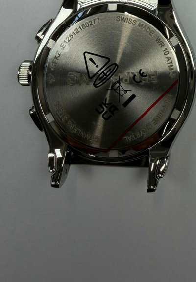 Parte trasera de reloj suizo de acero inoxidable que muestra resistencia al agua 10 ATM, marcas CE, UKCA, símbolos de advertencia y número de serie.