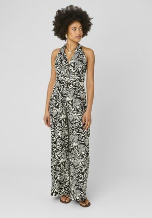 Femme avec une coiffure afro portant une combinaison à fleurs noire et blanche avec col halter et des sandales marron, debout et regardant sur le côté.