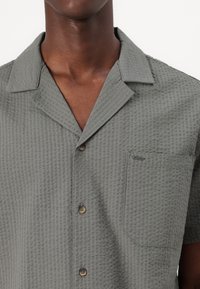 Camisa gris de manga corta con un patrón texturizado, cuello y un bolsillo. Presenta cierres de botones en un tono contrastante.