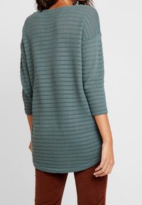 Maglione teal dalla vestibilità ampia con texture a coste e maniche a tre quarti, dotato di un orlo curvo e un scollo rilassato, abbinato a pantaloni color ruggine.