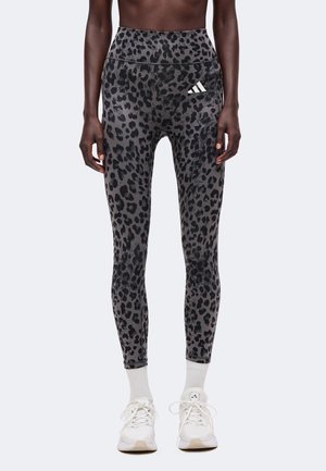 Kvinde iført højtaljede mørkegrå leggings med leopardprint, hvide sokker og hvide sportssko, der står mod en ensfarvet baggrund.