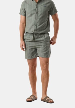 Mann trägt ein passendes, strukturiertes kurzärmeliges Hemd mit Knöpfen und Shorts in Olivgrün sowie braune Sandalen mit Schnalle.