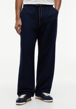 Marineblaue, weit geschnittene Hose mit Kordelzug in der Taille, getragen mit marineblauen und weißen Freizeitschuhen an einer stehenden Person.