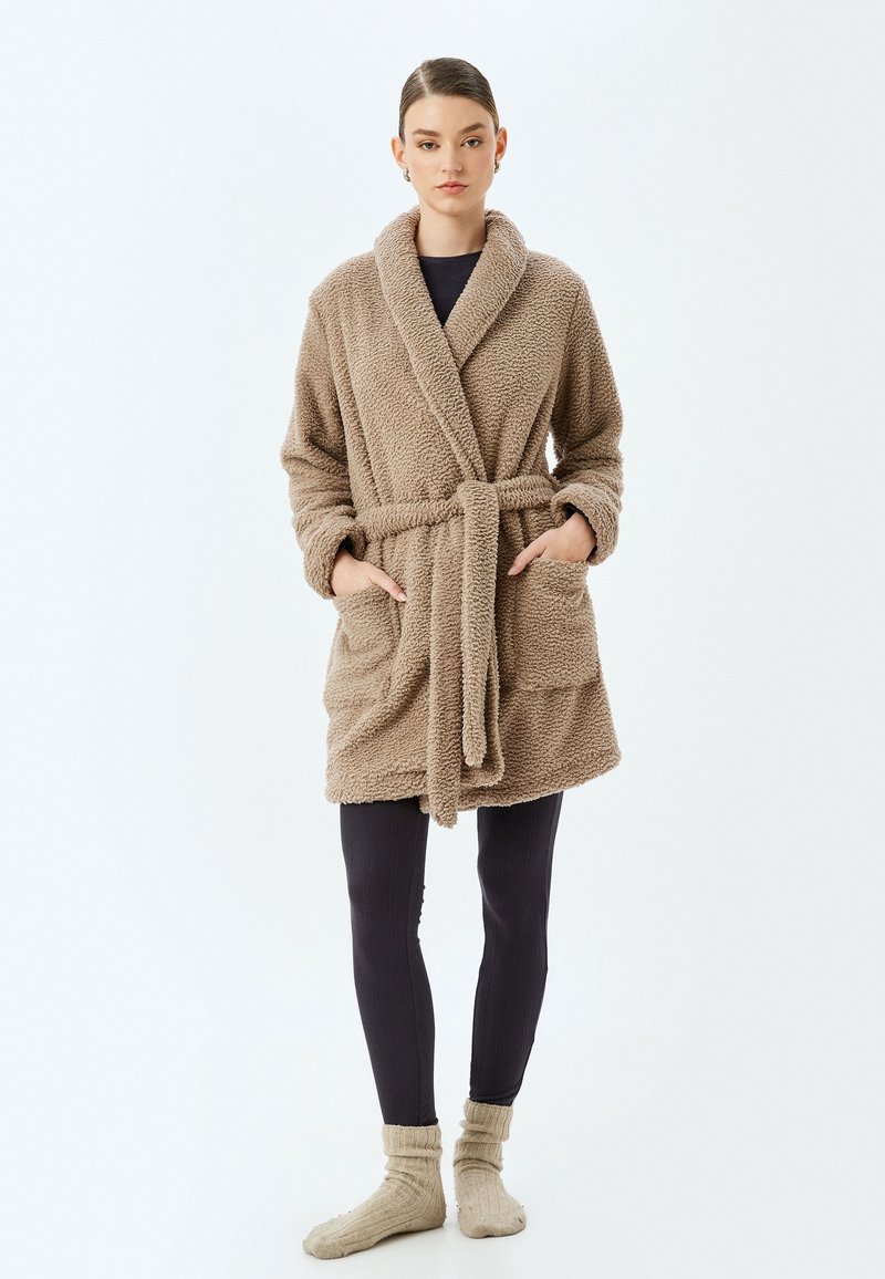 Accappatoio teddy beige e soffice con colletto a scialle e vita con cintura, dotato di due tasche frontali e maniche rilassate, abbinato a leggings scuri.