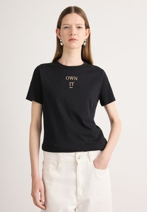 T-shirt en coton noir à manches courtes avec le texte "POSSEDEZ-LE" brodé en or au centre. Le modèle porte un pantalon blanc taille haute.