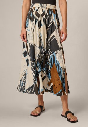 Woman wearing a pleated skirt with abstract blue, black, white, and brown patterns, paired with black open-toe sandals with gold accents.