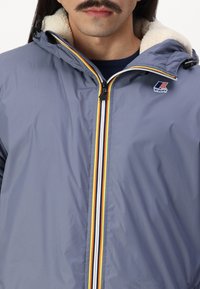 Giacca blu con cappuccio, foderata in morbido crema, dotata di una cerniera centrale e strisce multicolori lungo la zip, con un logo sul petto.