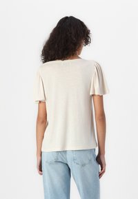 Bežinis trumpas rankovėmis viršus su suspaudžiamosiomis rankovėmis, apvaliu kaklu ir minkšta tekstūra, derinamas su šviesiai mėlynomis džinsinomis kelnėmis.