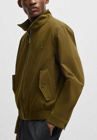 Olive green lightweight jacket with a high collar, front zipper, elastic hem, and two angled pockets featuring button closures.
