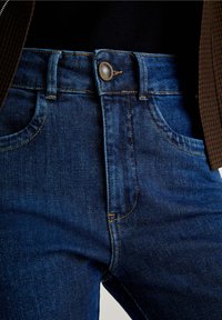 Blue denim jeans feature a classic five-pocket design, zip and button closure, with visible stitching and slight fading near the pockets.