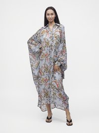 Femme portant une robe longue, ample et fleurie avec de larges manches et des sandales noires à enfiler, debout avec une main sur la hanche.