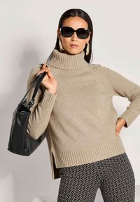 Frau in beigem Rollkragenpullover, schwarzen gemusterten Hosen, großen goldenen Creolen, schwarzen Sonnenbrillen und hält eine schwarze Handtasche.